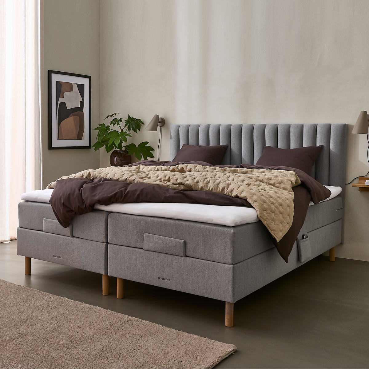 Lyst soveværelse med grå kontinentalseng, beige quiltet plaid og mørkebrunt sengetøj, væglamper, plante og vægkunst ved et stort vindue med lyse gardiner.