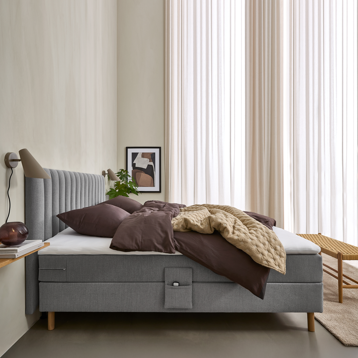 Lyst soveværelse med grå polstret seng, brunt sengetøj og beige quilt; lyse gardiner, væglamper, lille hylde med bøger og glaslampe, plante og kunst.