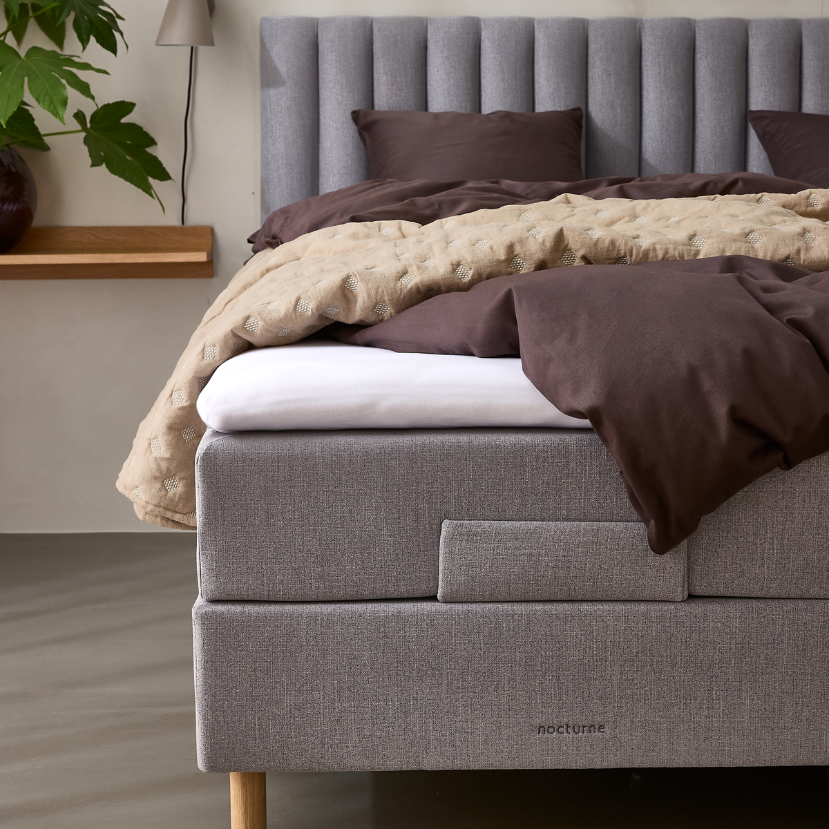 Nærbillede af en grå polstret seng med kanalsyet gavl, brunt sengetøj og beige quilt samt træben, med mærket nocturne.