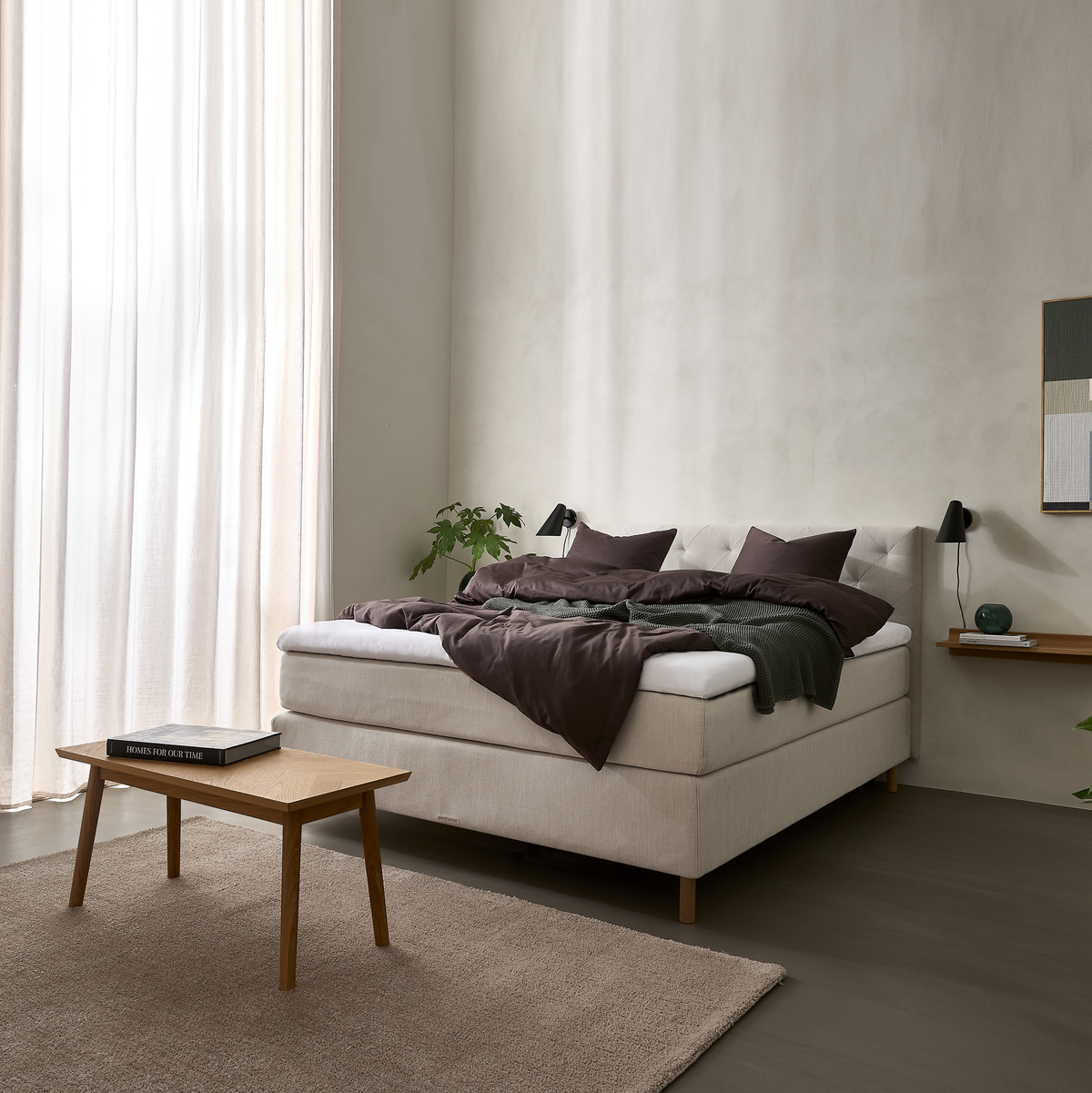 Lyst soveværelse med beige kontinentalseng, mørkebrunt sengetøj og grønt plaid, sorte væglamper, lille træbord og neutral tæppe i blødt dagslys.