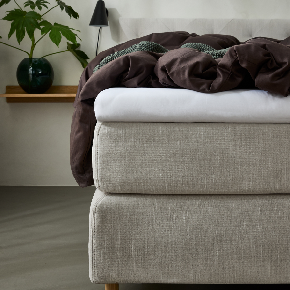 Beige polstret seng med hvidt lagen, brun dyne og grøn strikket plaid; sort væglampe og træhylde med grøn plante i glasvase.
