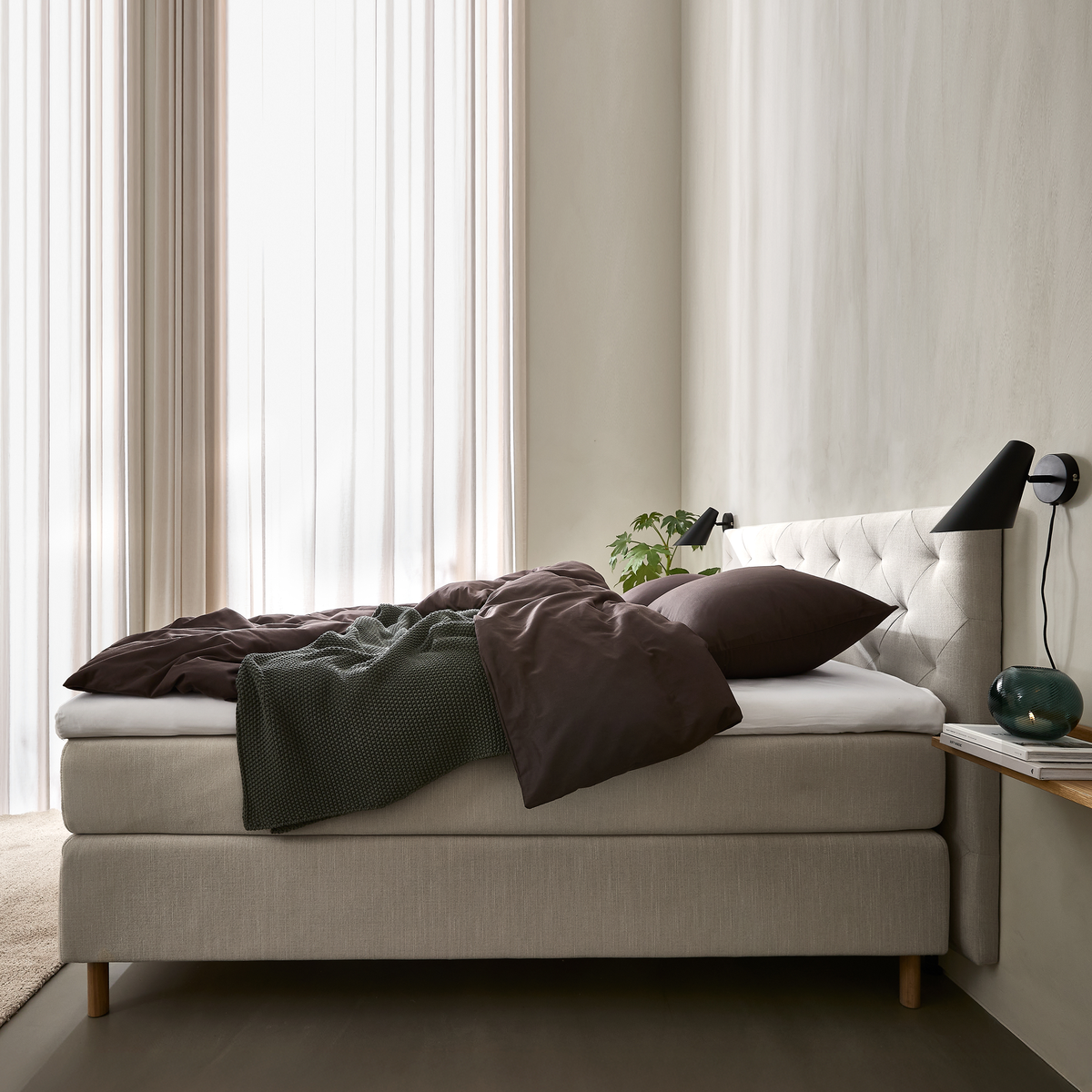 Lyst soveværelse med beige kontinentalseng og polstret sengegavl, mørkebrunt sengetøj og grøn plaid, sorte væglamper, lette gardiner samt lille hylde med glasvase og bøger.