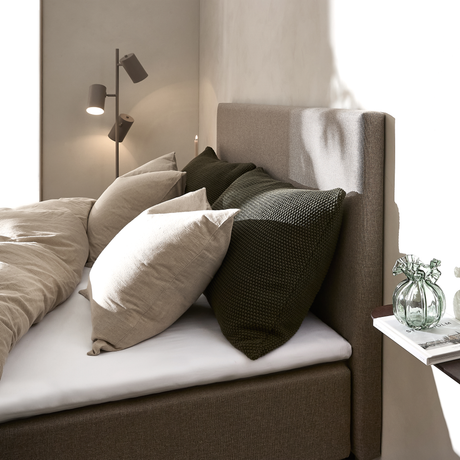 Nærbillede af en redt seng med beige og mørkegrønne puder, polstret brun sengegavl, en gulvlampe samt en lille hylde med glasvase og bog i blødt dagslys.