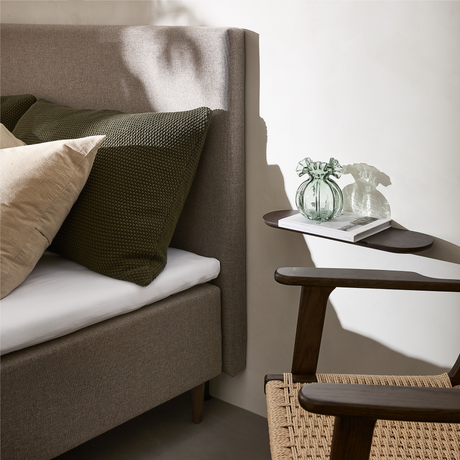 Soveværelseshjørne med beige polstret seng, grønne og beige puder, lille væghylde med grøn glasvase og bog samt stol i mørkt træ med flet-sæde.