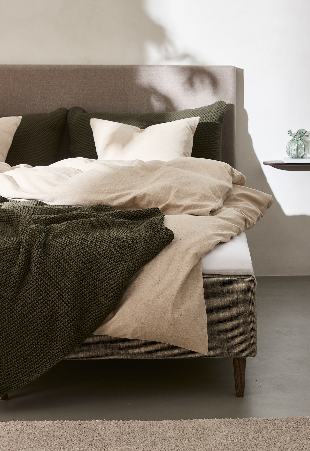 Beige linsengetøj med mørkegrønt strikket plaid på en polstret seng i et lyst, minimalistisk soveværelse.