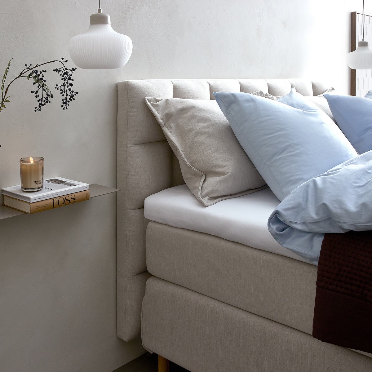 Lyst og roligt soveværelsesmiljø med beige kontinentalseng, lyseblåt sengetøj, puder, et mørkt strikket plaid og en hvid pendellampe; på en væghylde står et lys og bøger, hvoraf en har titlen FOSS.