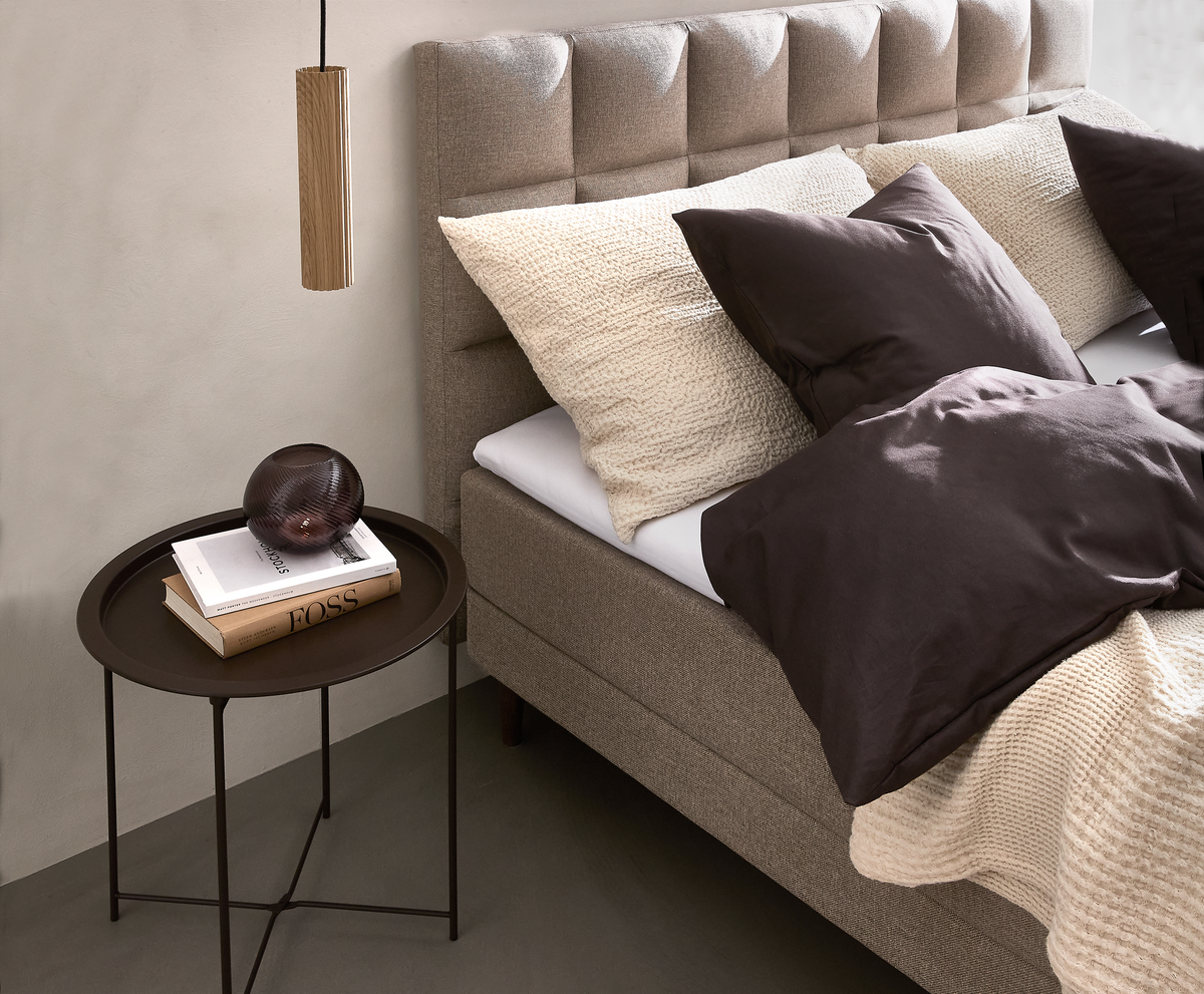 Soveværelse med beige polstret seng, mørkebrune og cremefarvede puder, strikket plaid, rundt sort sengebord med glasdekoration og bøger – en med titlen FOSS – samt hængende træpendel.