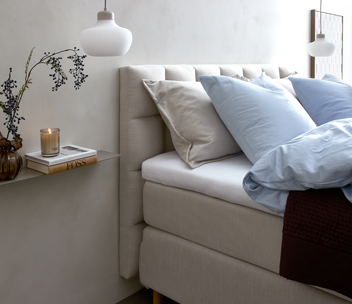 Beige polstret seng med lyseblåt sengetøj og puder, hvid glaspendel, smal hylde med vase med gren, tændt duftlys og bog med titlen FOSS.