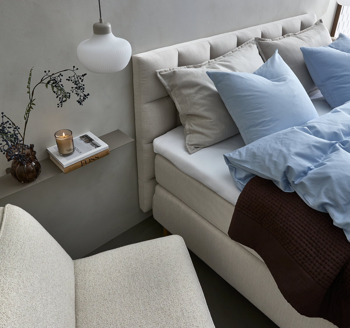Lyst og roligt soveværelsesmiljø med beige polstret seng, lyseblåt sengetøj med grå puder, mørkebrun plaid ved fodenden, en hvid pendellampe og en smal hylde med vase, tændt duftlys og en bog med titlen HOSS.
