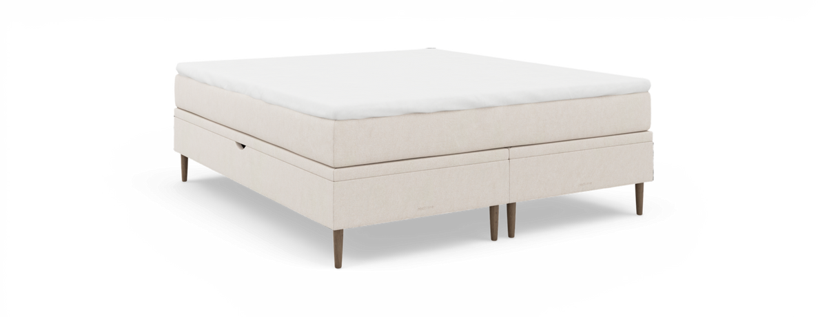 Beige kontinentalseng med hvid topmadras, fritstående mod sort baggrund.