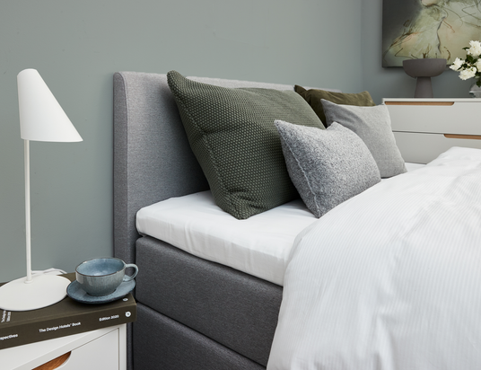 Nærbillede af en grå polstret seng med hvidt sengetøj og grønne/grå pyntepuder, et natbord med hvid bordlampe, kop og “The Design Hotels Book, Edition 2020”; i baggrunden en kommode med vase og hvide blomster samt vægkunst.