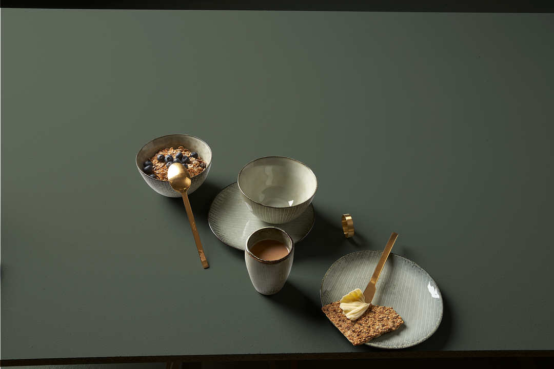 Minimal morgenopdækning med keramikskåle, kop og tallerkener, gylden ske og smørkniv, granola med blåbær og knækbrød med smør på en mørkegrøn overflade.
