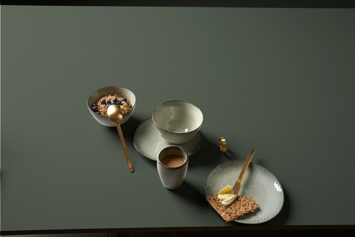 Minimal morgenopdækning med keramikskåle, kop og tallerkener, gylden ske og smørkniv, granola med blåbær og knækbrød med smør på en mørkegrøn overflade.