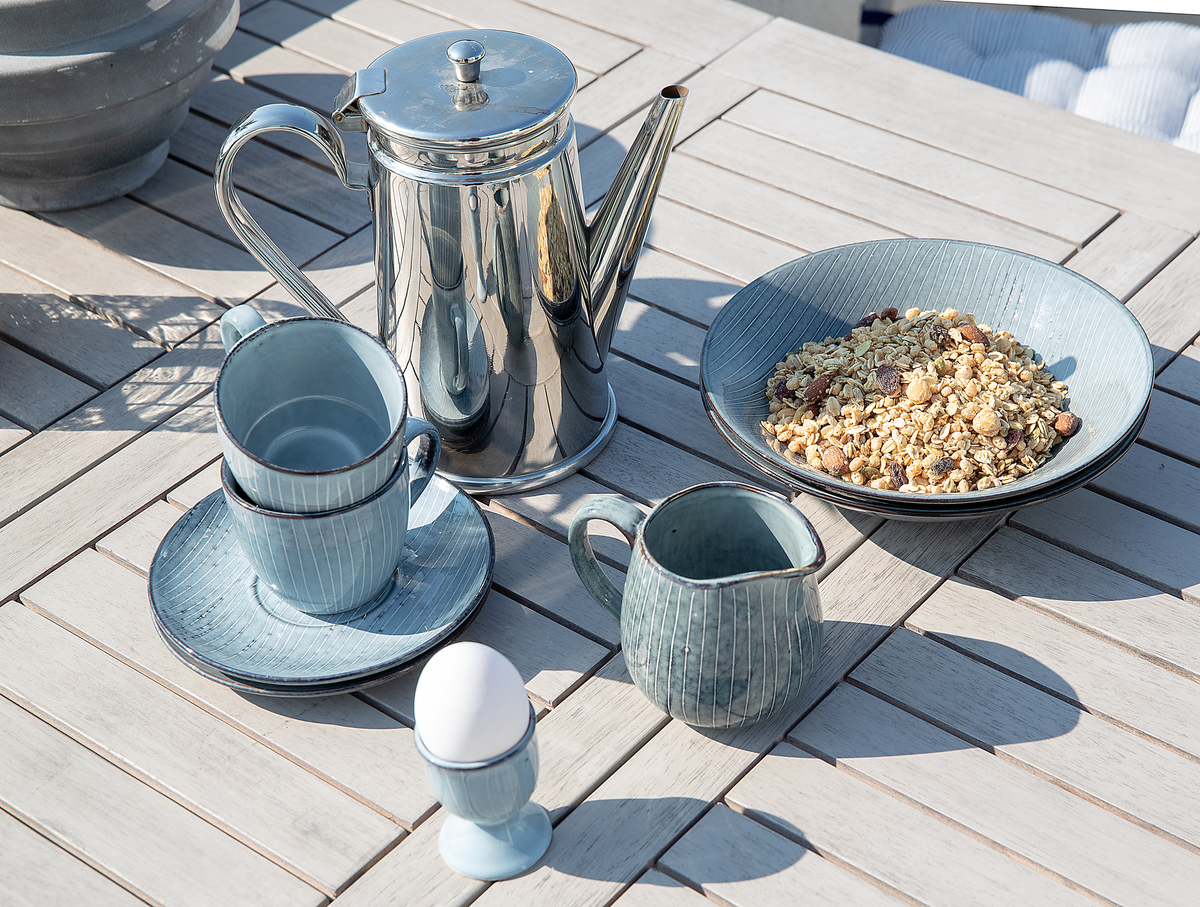 Udendørs morgenmad med rustfri kaffekande, blå kop med underkop, lille kande, æg i æggebæger og en skål med müsli på et havebord i sollys.