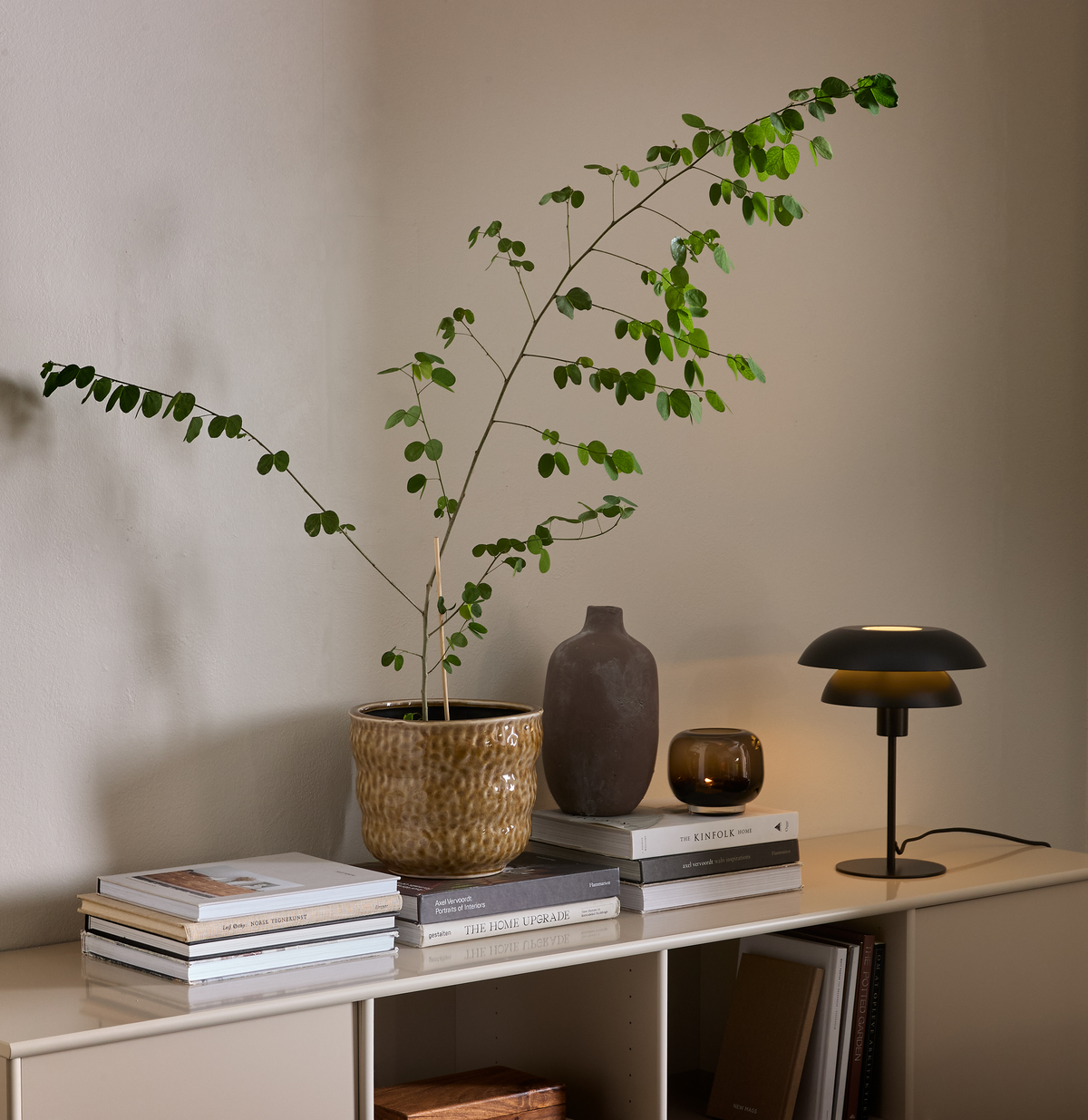 Beigefarvet reol med stakke bøger (The Home Upgrade og The Kinfolk Home), en grøn potteplante, brun keramikvase, lille ravfarvet lysestage og sort bordlampe mod en lys væg.