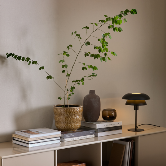 Beigefarvet skænk med grøn potteplante, keramikvase, tændt lysglas og sort bordlampe oven på stakke af bøger som The Kinfolk Home og The Home Upgrade mod en neutral væg.