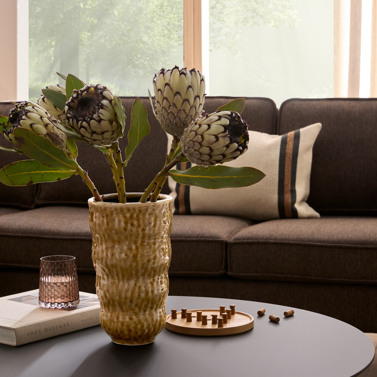 Nærbillede af en stue: keramikvase med protea på sofabordet, riflet glas, bogen ”Soft Minimal” og et lille træspil foran en brun sofa med stribet pude ved et lyst vindue.