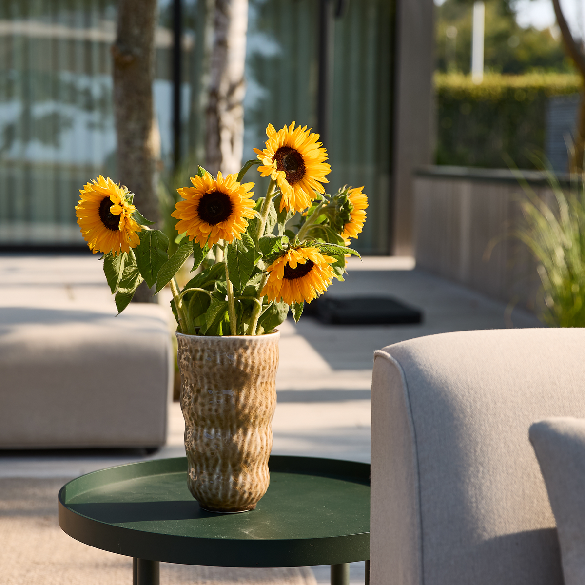 Solsikker i en keramisk vase på et grønt sidebord på en solrig terrasse.