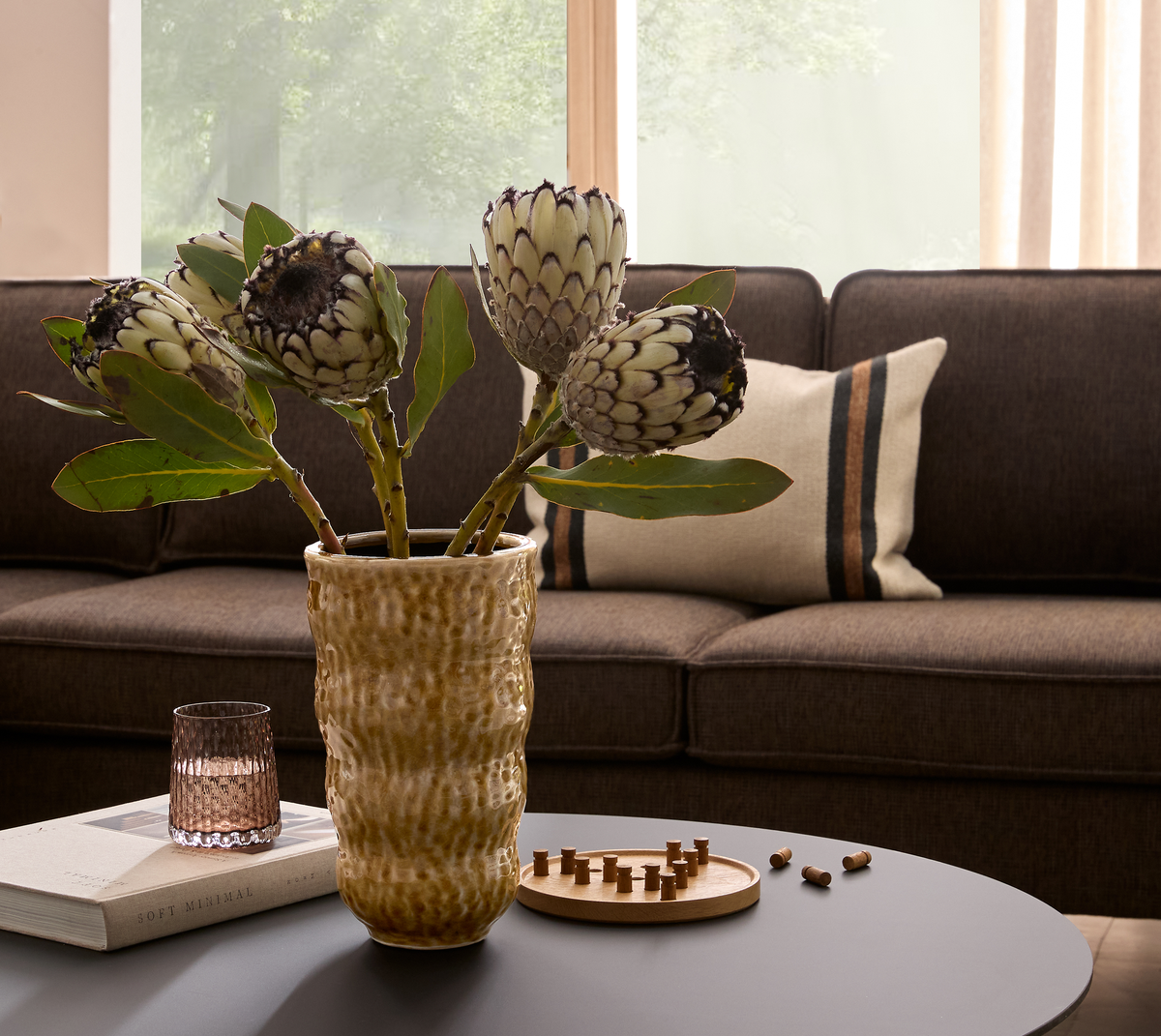 Protea-blomster i en rillet beige vase på et sofabord med glas, bogen Soft Minimal og et lille træspil; brun sofa med stribet pude og vindueslys i baggrunden.