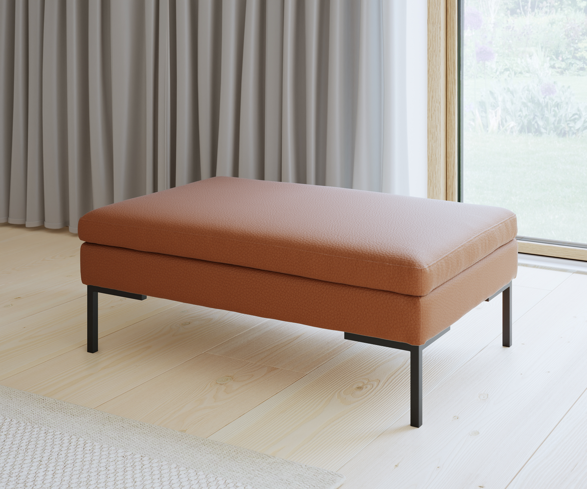 Lav ottoman med terrakottafarvet polstring og sorte metalben i en lys stue med store vinduer og grå gardiner.
