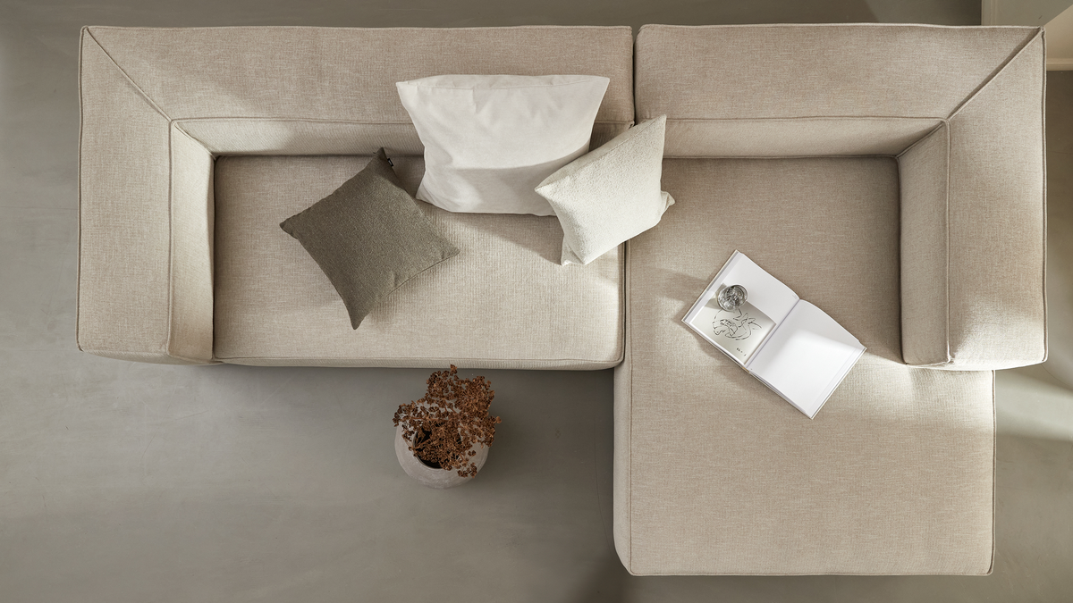 Beige modulsofa set oppefra med puder, en bakke med åbnet bog og glas samt en potteplante på gulvet.