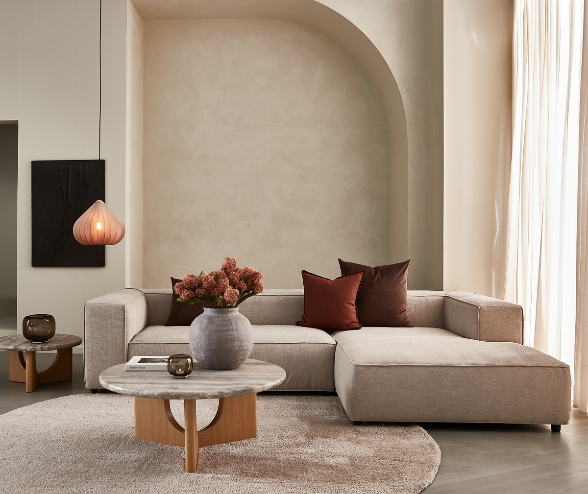 Beige hjørnesofa i stue med rundt sofabord i træ og sten, vase med rosabrune blomster, to mørke puder, hængende pendellampe og lyse gardiner i blødt dagslys.