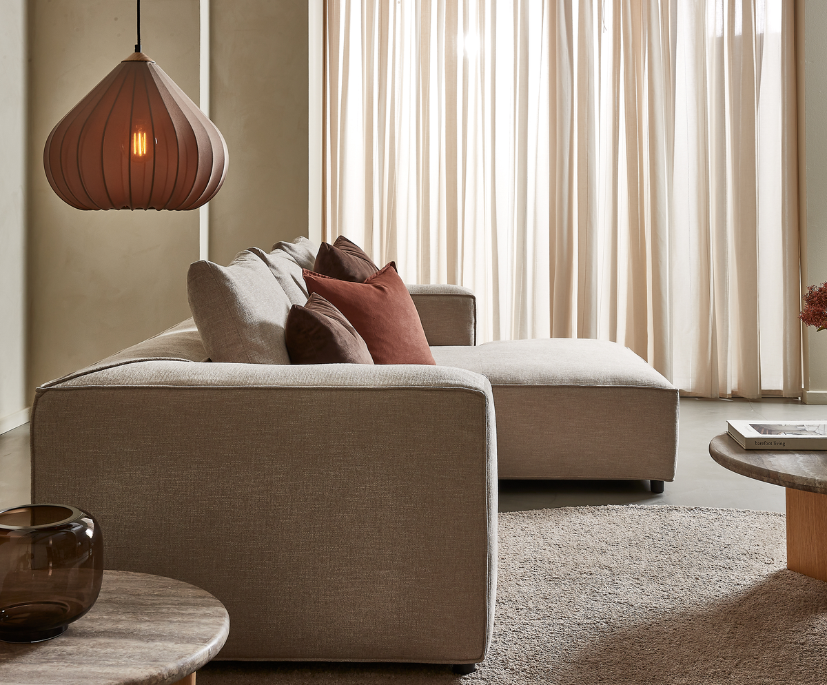 Beige hjørnesofa med rust- og brune puder, rundt sofabord med glasvase, hængende pendel og lyse, lette gardiner i en rolig stue.