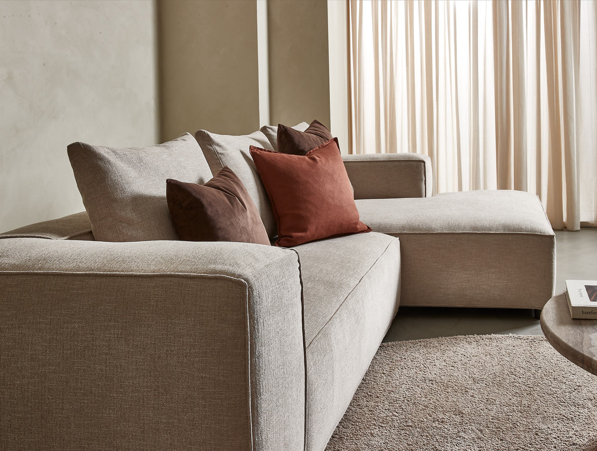 Beige hjørnesofa med chaiselong og rustfarvede puder i en lys stue med lyse gardiner og tæppe.