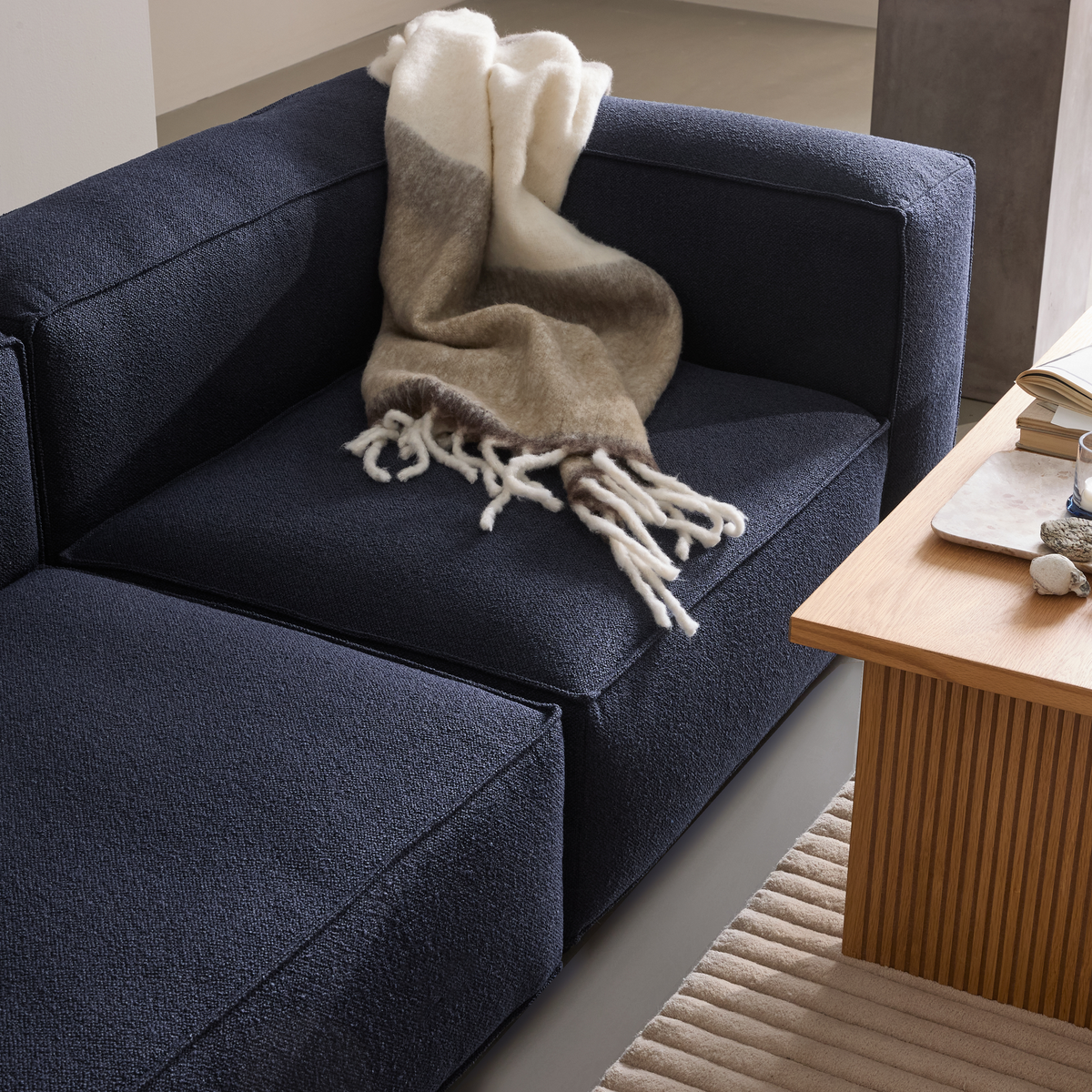 Mørkeblå modulsofa i bouclé med blød uldplaid; ved siden af står et lyst egetræsbord med bakke, bog og glas på et beige, riflet tæppe.