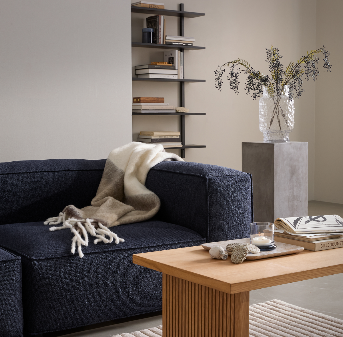 Stilren stue med mørkeblå sofa, beige plaid, træbord med lys, sten og bøger, hvoraf en hedder BILLEDKUNST; væghylde og glasvase med grene på sokkel.