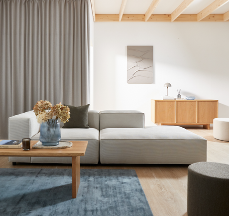 Lyst skandinavisk stue med lysgrå modulsofa, træsofabord med vase med tørrede blomster, bogen ”Classic Houses” og lys, blå tæppe samt rillet træskænk med lille bordlampe mod hvid væg.