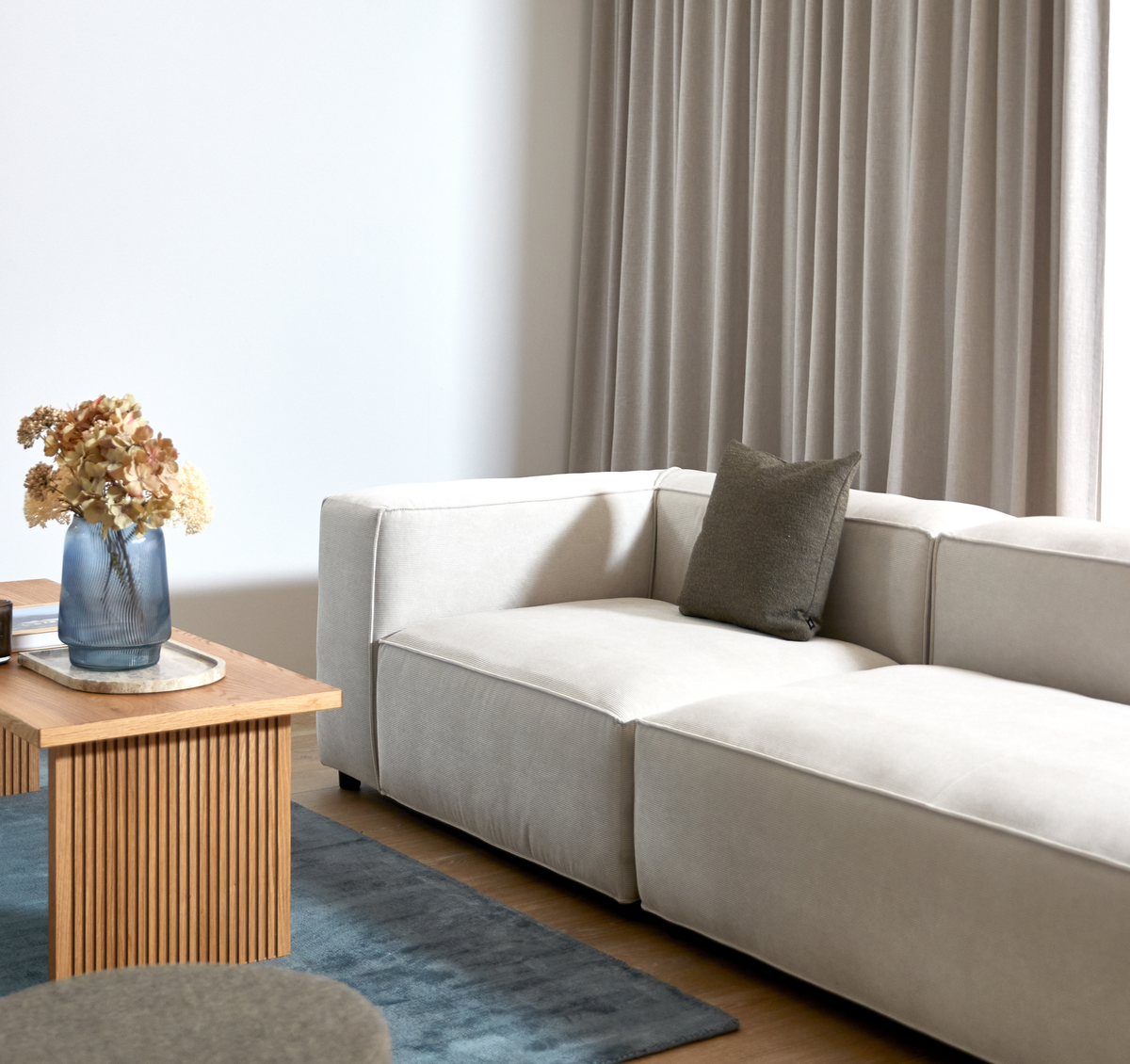 Lyst stue med beige modulsofa, grå pude, rillet sofabord i træ med blå glasvase med tørrede blomster, gråt tæppe og beige gardiner.