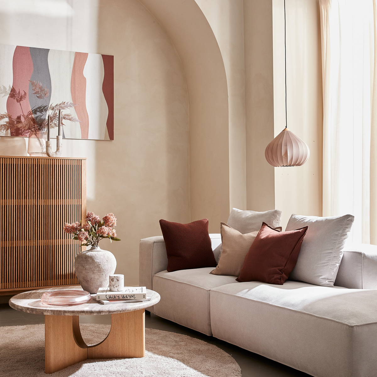 Lys skandinavisk stue med beige sofa, rust- og sandfarvede puder, rundt træ/sten-sofabord med vase, lys og bogen CHLOE SEVIGNY; ribbet skænk, abstrakt vægkunst og rosa pendellampe i blødt dagslys ved lette gardiner.