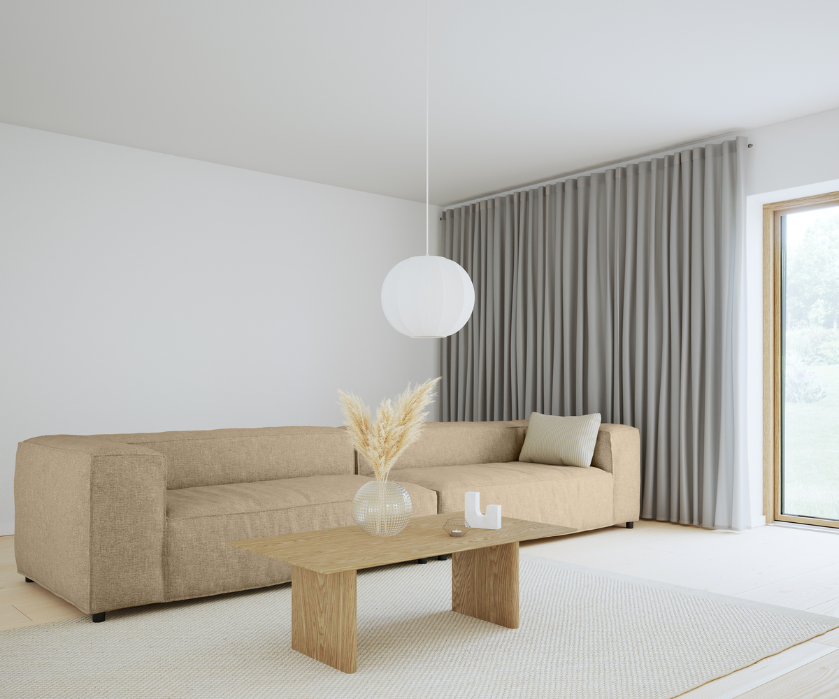 Lyst skandinavisk stue med beige hjørnesofa, lyst sofabord i træ, rund hvid pendel, grå gulvlange gardiner og en vase med tørret græs ved et stort vindue.