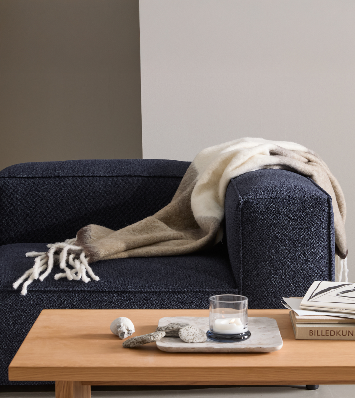 Mørkeblå sofa med uldplaid, lavt sofabord i træ med stentallerken, sten og tændt fyrfadslys i glas; bøger med øverste titel ”BILLEDKUN...” i en lys, minimalistisk stue.