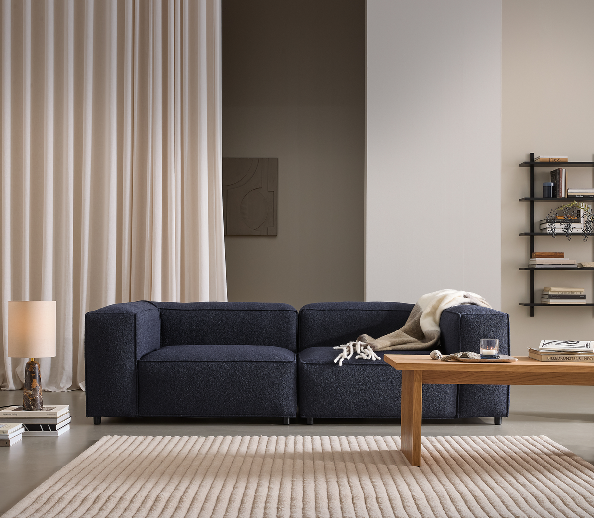 Mørkeblå modulsofa i moderne stue med lyst ribbet tæppe, træsofabord, beige gardiner, væghylder, bordlampe og en plaid over sofaen.