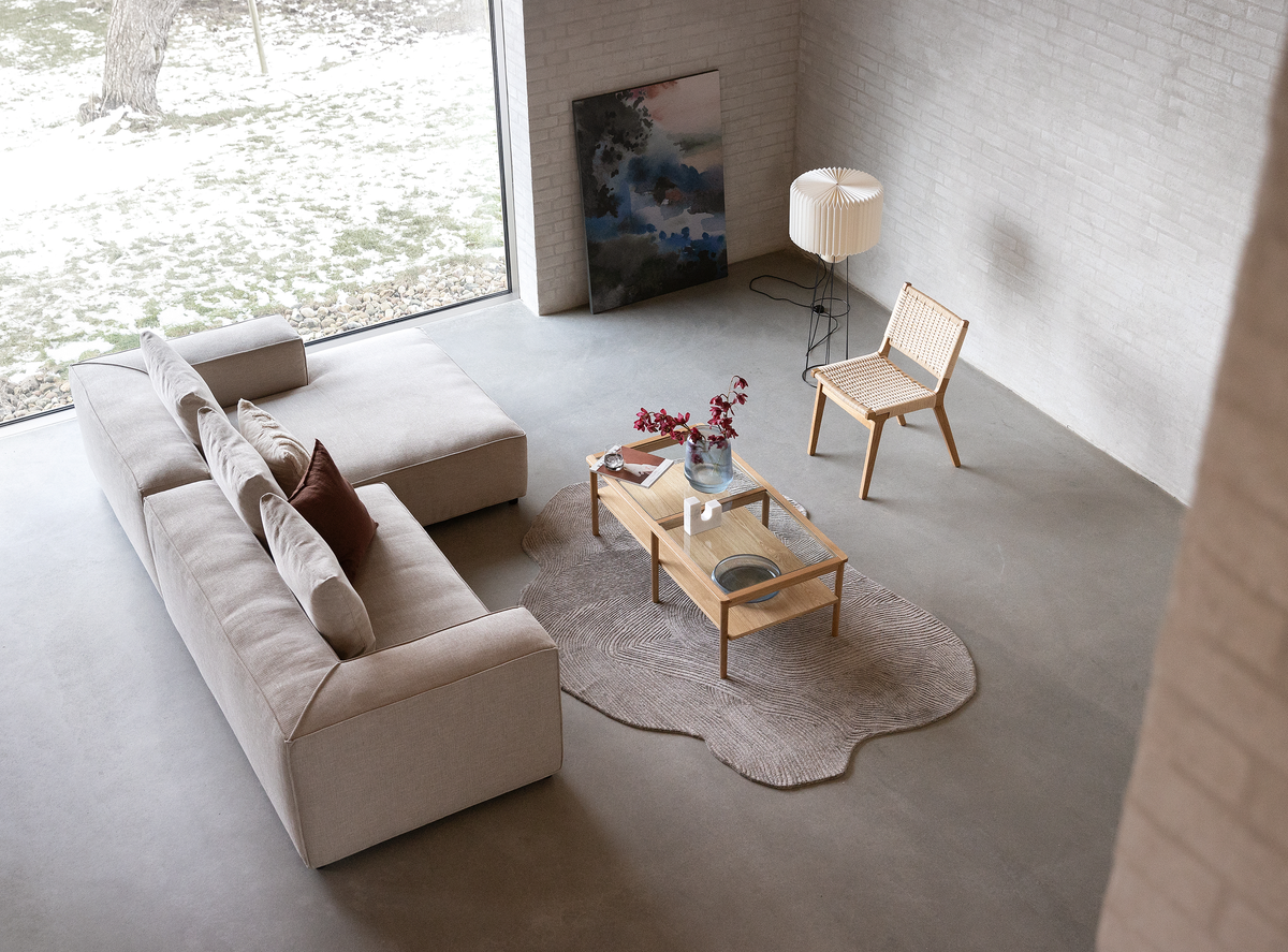 Lyst stue med beige sofa, fletstol i træ og sofabord i træ på organisk formet tæppe, gulvlampe og maleri ved stort vindue.