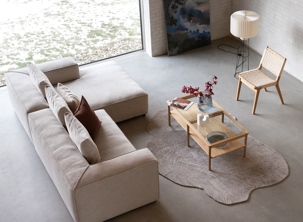 Lys skandinavisk stue med beige modulsofa, træ-sofabord med glas, organisk formet tæppe og plisseret gulvlampe ved stort vindue.