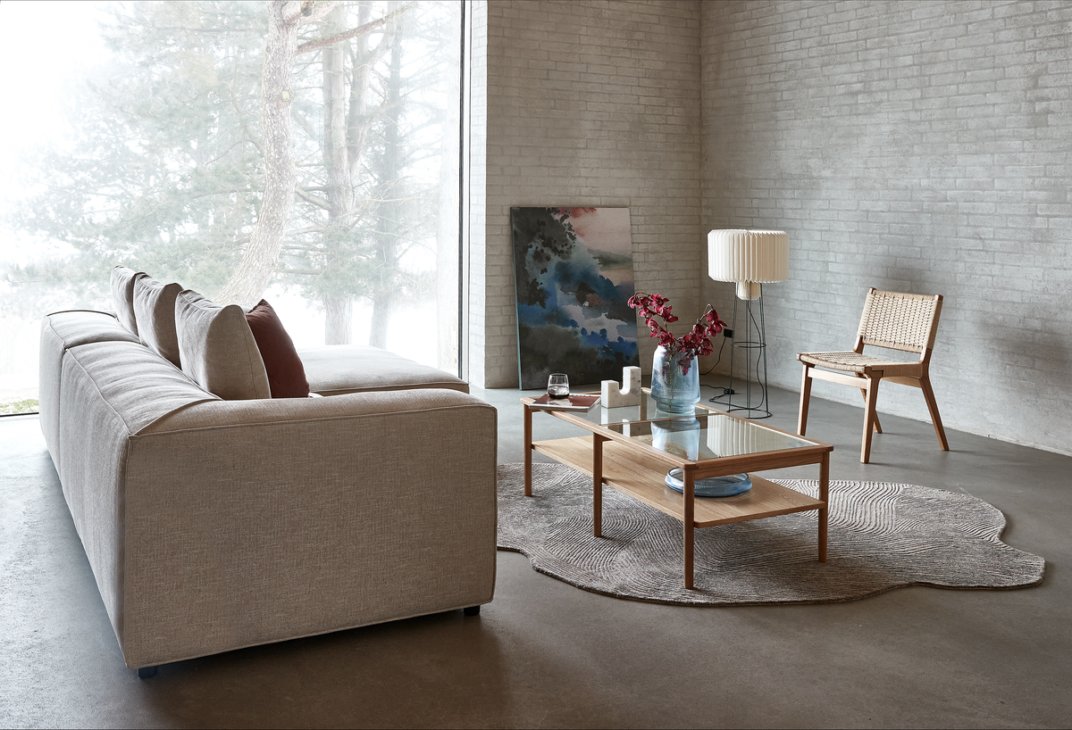 Lyst skandinavisk stue med beige sofa, glasbord i træ, fletstol, gulvlampe, organisk gråt tæppe og et maleri, der læner sig mod en lys murstensvæg ved et stort vindue.