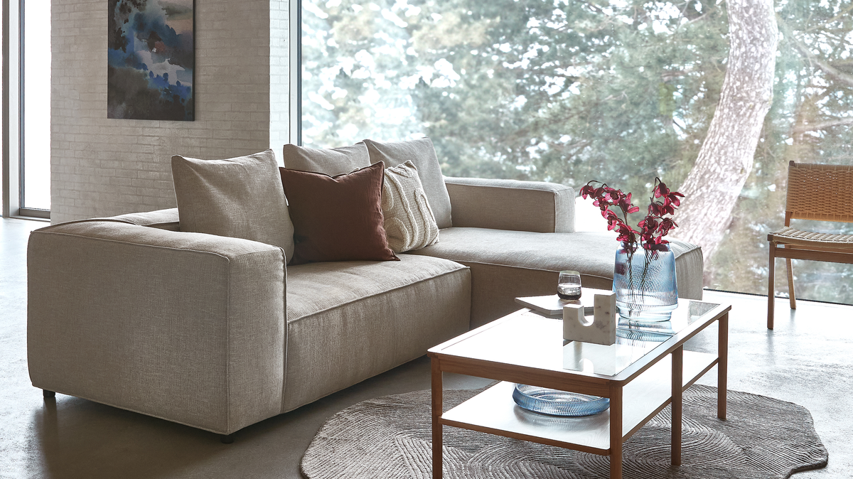 Lys stue med beige hjørnesofa, puder, glasbord i træ og blå vase med pink blomster ved store vinduer mod træer.