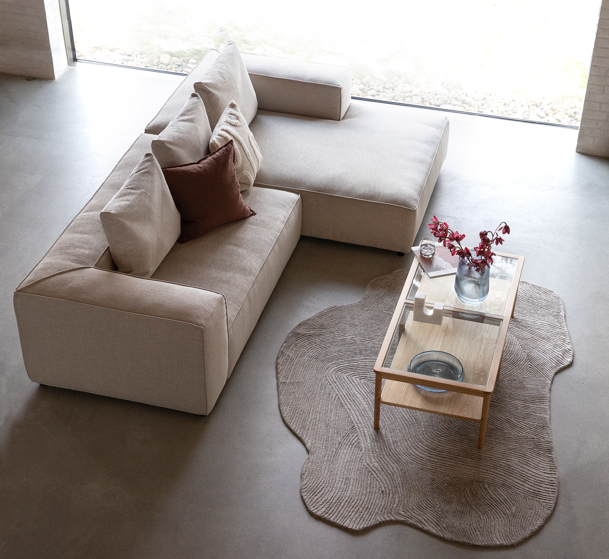 Top-down visning af lyst stue med beige modulsofa, glas/træ-sofabord med vase og røde grene på et organisk formet beige tæppe.