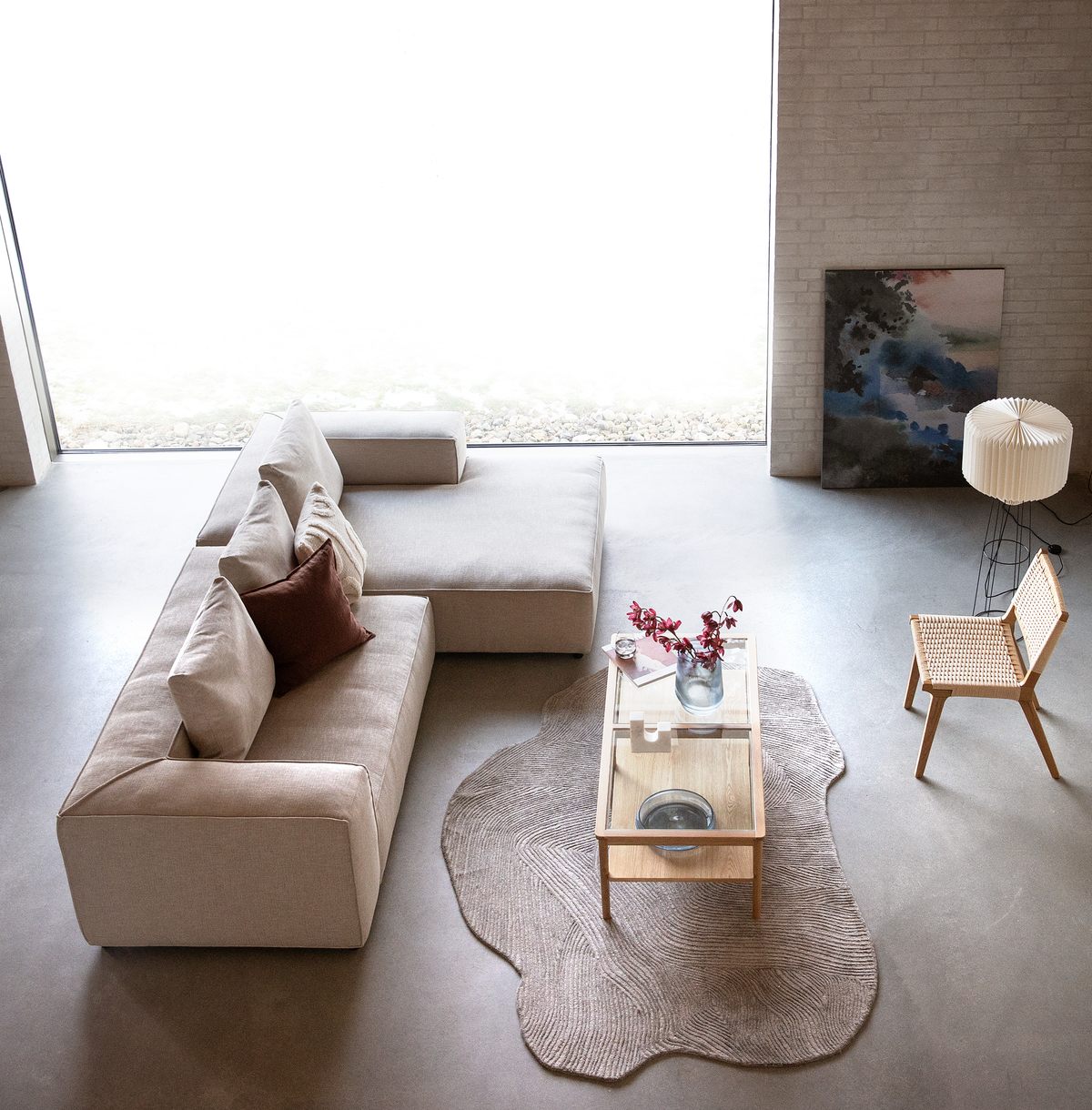 Lys skandinavisk stue med beige modulsofa, glasbord i træ på organisk lys tæppe, fletstol og plisseret gulvlampe ved stort vindue; vase med røde grene og maleri op ad væggen.