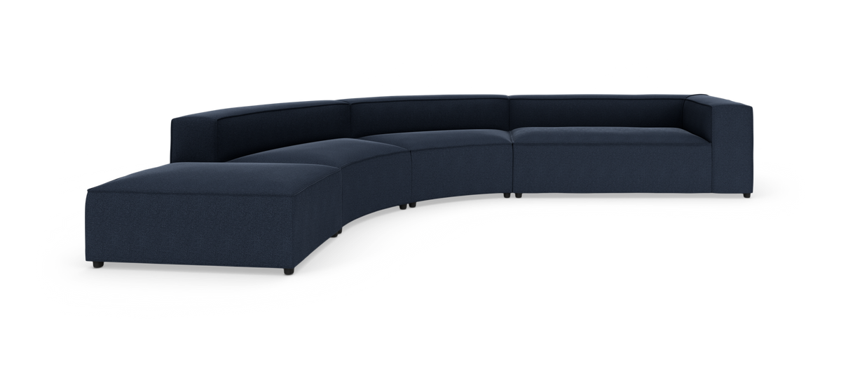 Modulær, buet sofa i mørkeblå med ottoman i venstre ende på hvid baggrund.