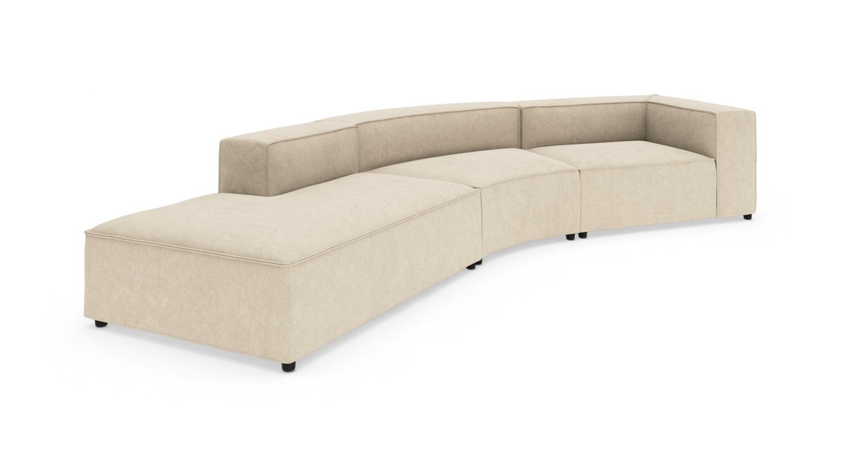 Beige modulsofa i buet L‑form med chaiselong til venstre, lavt ryglæn og blokarmlæn på sorte fødder, fritlagt mod hvid baggrund.
