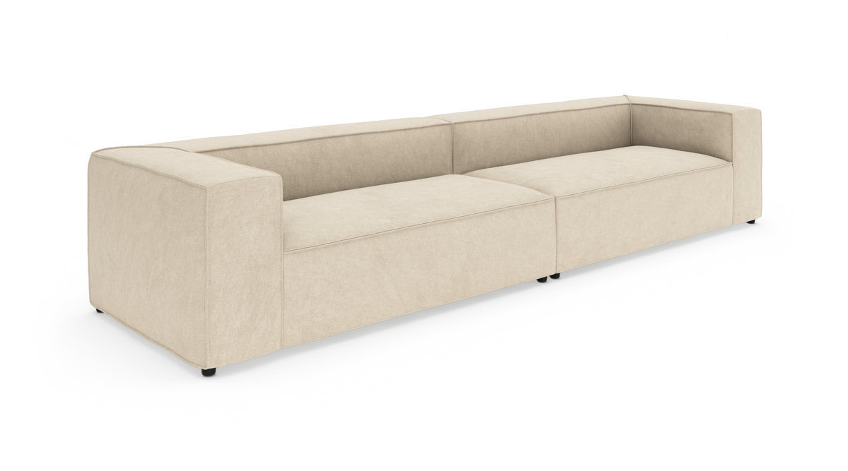Beige, lav modulsofa med brede armlæn, set skråt forfra på hvid baggrund.