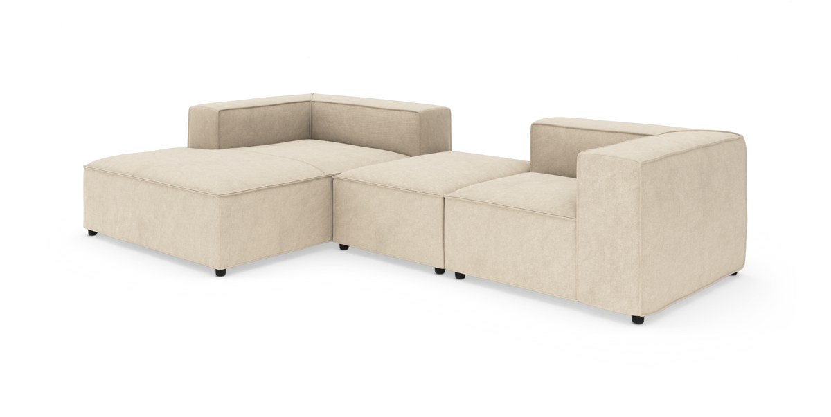 Beige modulsofa i L‑form med bred chaiselong og lave sorte ben, frilagt på hvid baggrund.