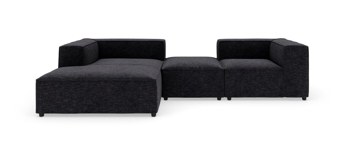 Modulær mørkegrå hjørnesofa med chaiselong til venstre og en midterpuf, fritstående på hvid baggrund.