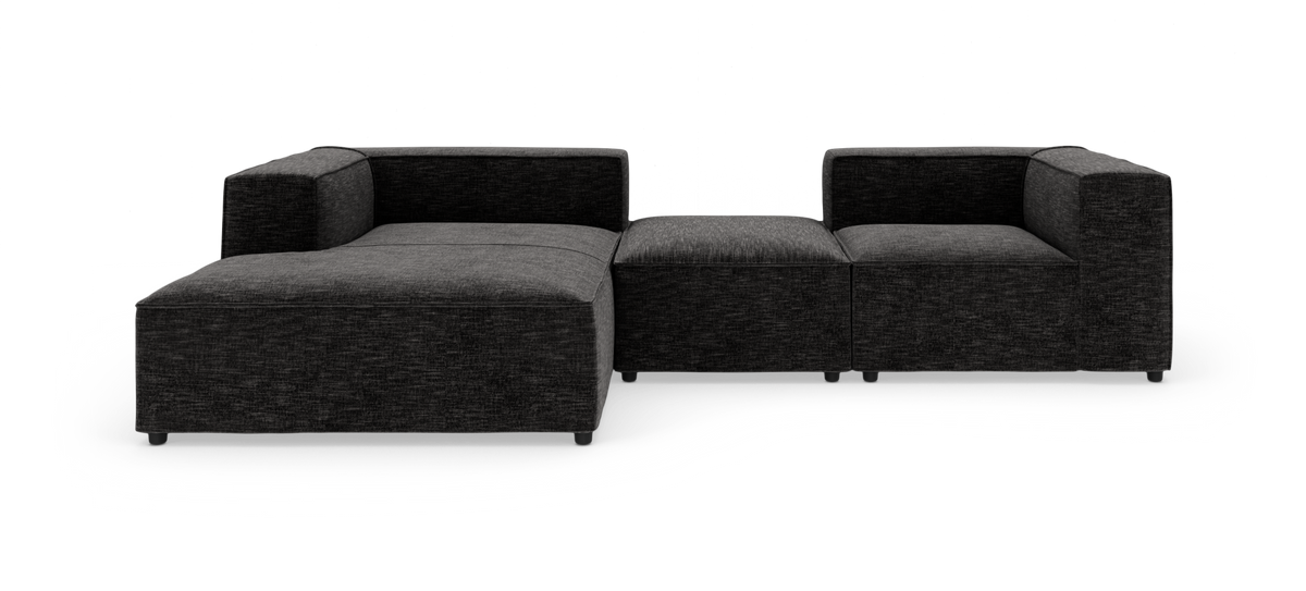 Modulær mørkegrå hjørnesofa med chaiselong til venstre og en midterpuf, frilagt på hvid baggrund.