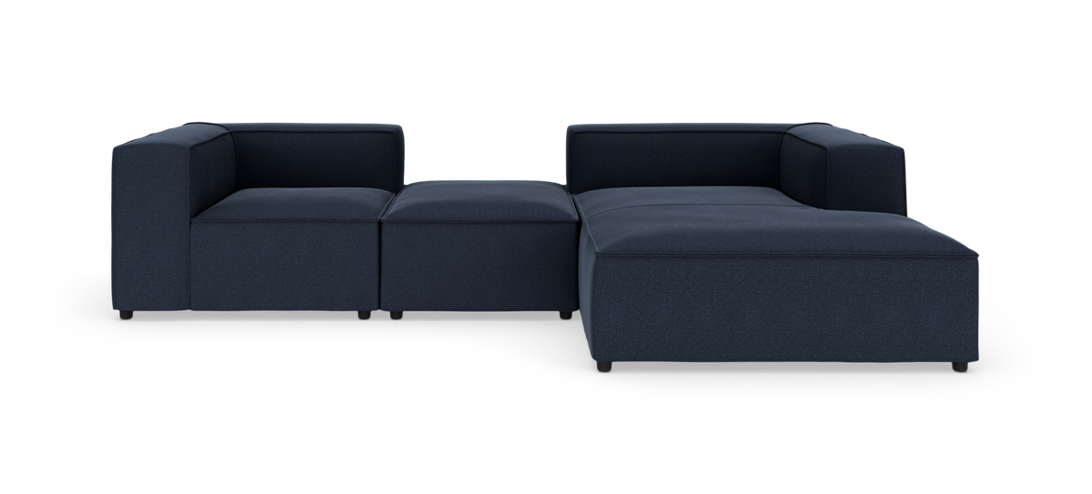 Modulsofa i mørkeblåt stof, L‑form med bred chaiselong og lave armlæn, produktfoto på hvid baggrund.