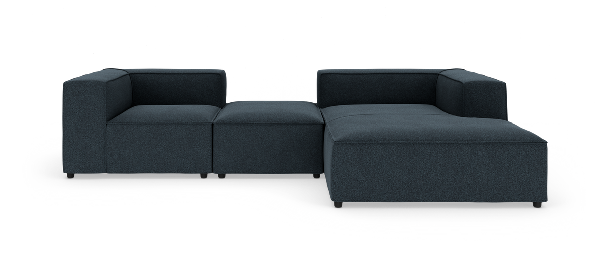 Modulsofa i mørkblåt stof, L‑form med lav ryg og bred chaiselong på hvid studiobaggrund.